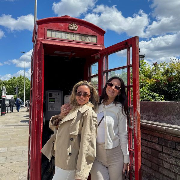 Two women smiling in front of a classic red British telephone booth on a sunny day, with blue sky and fluffy clouds in the background.