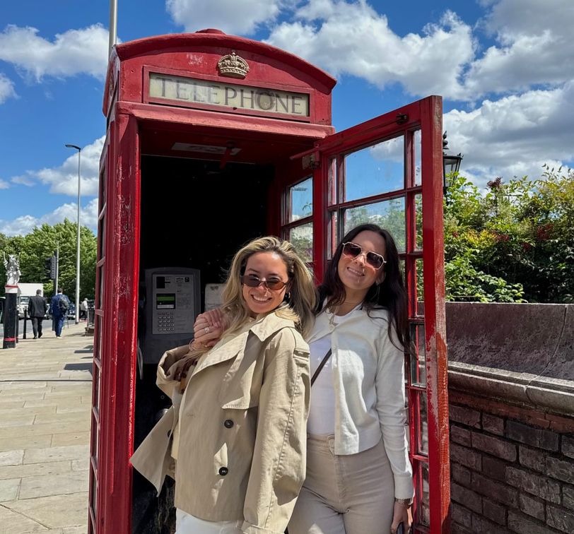 Two women smiling in front of a classic red British telephone booth on a sunny day, with blue sky and fluffy clouds in the background.