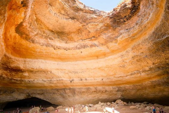 A sunlit cave with a sandy beach inside, surrounded by rocky walls. People are standing and kayaks are nearby. An opening above lets in sunlight.