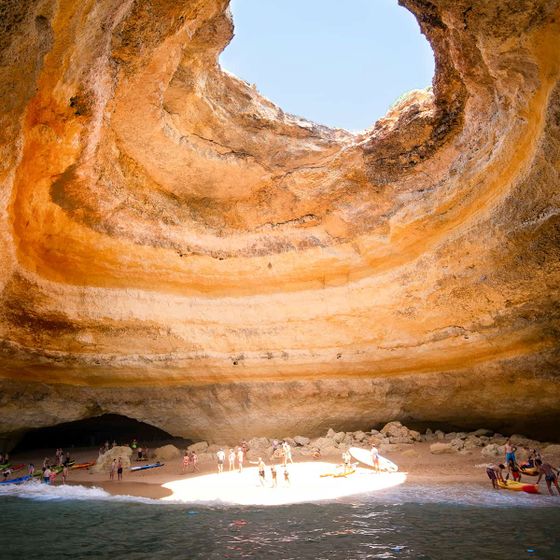 Tourists and kayaks inside a sunlit coastal cave with layered rock formations and a visible opening at the top.