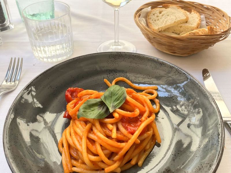 A plate of spaghetti with tomato sauce and basil, a glass of white wine, bread in a basket, and a water glass on a white tablecloth.