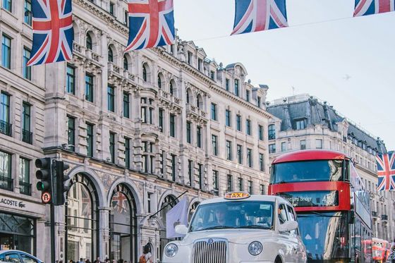 Street scene in London with a classic white taxi and red double-decker bus, surrounded by Union Jack flags and historic architecture.