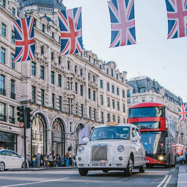 Street scene in London with a classic white taxi and red double-decker bus, surrounded by Union Jack flags and historic architecture.