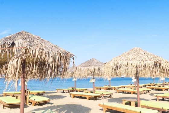 Sandy beach with wooden sun loungers under thatched umbrellas, beside clear blue waters under a bright blue sky.
