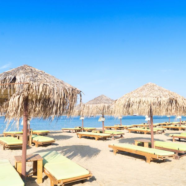 Sandy beach with wooden sun loungers under thatched umbrellas, beside clear blue waters under a bright blue sky.