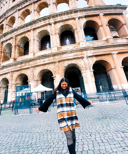 A person in a plaid dress stands joyfully with arms outstretched in front of the Colosseum on a cobblestone street.