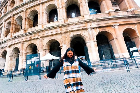 A person in a plaid dress stands joyfully with arms outstretched in front of the Colosseum on a cobblestone street.