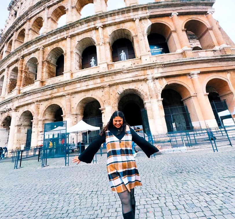 A person in a plaid dress stands joyfully with arms outstretched in front of the Colosseum on a cobblestone street.