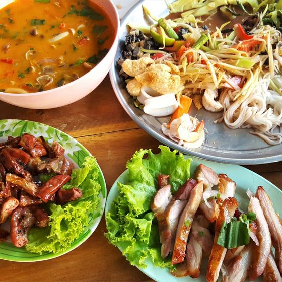 A colorful Thai meal with spicy soup, papaya salad, grilled meat on lettuce, and fresh vegetables on a wooden table.