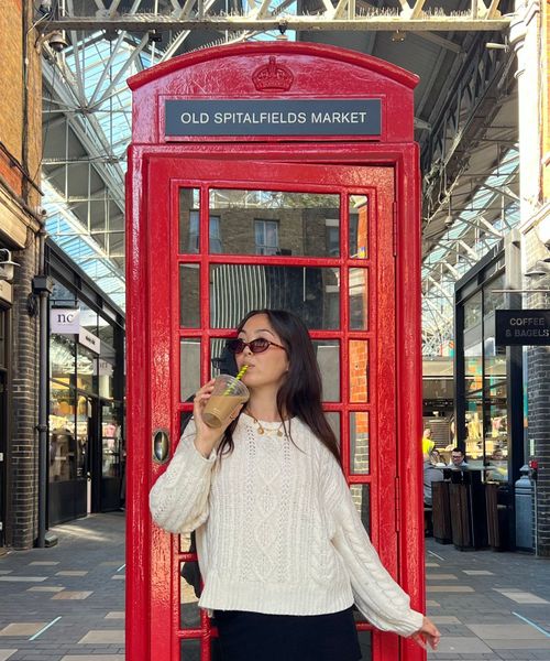 A person in sunglasses drinks from a cup in front of a red phone booth at Old Spitalfields Market, surrounded by brick buildings and glass roof.