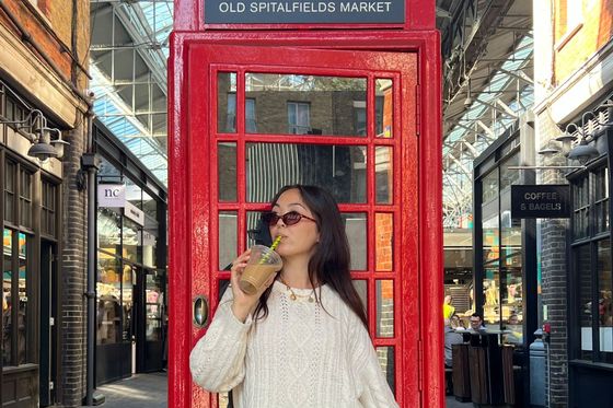 A person in sunglasses drinks from a cup in front of a red phone booth at Old Spitalfields Market, surrounded by brick buildings and glass roof.