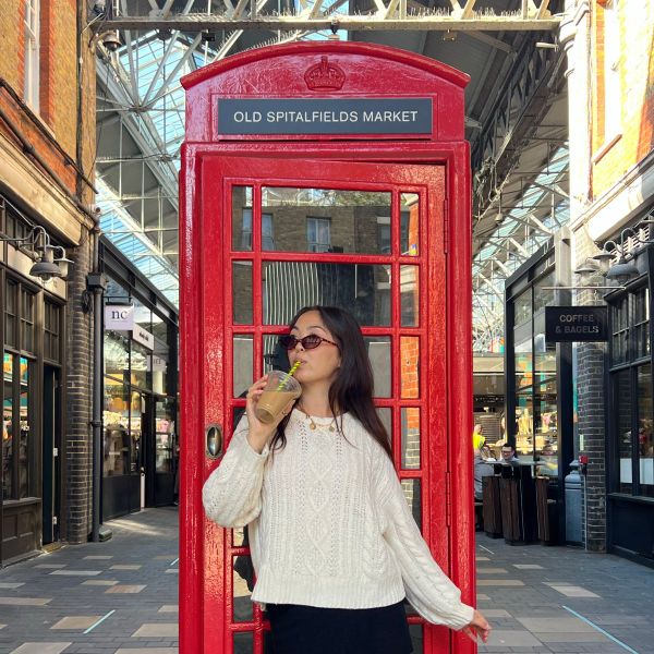 A person in sunglasses drinks from a cup in front of a red phone booth at Old Spitalfields Market, surrounded by brick buildings and glass roof.