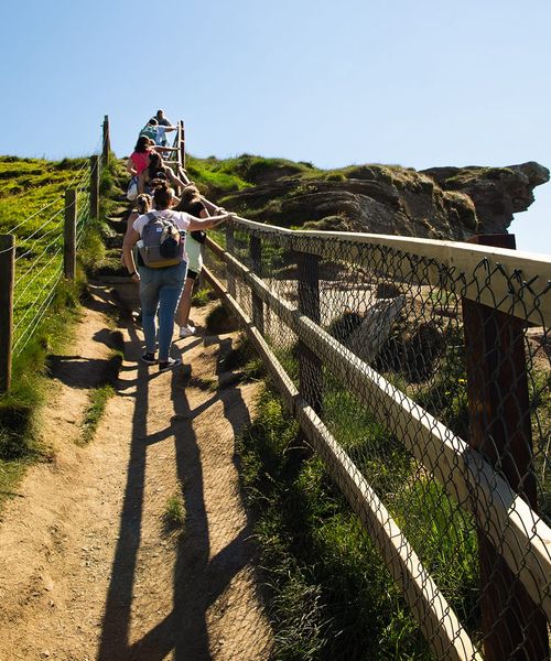 People walking up a narrow dirt path on a sunny day, flanked by grassy hills and a wooden fence, leading to a rocky cliff edge.