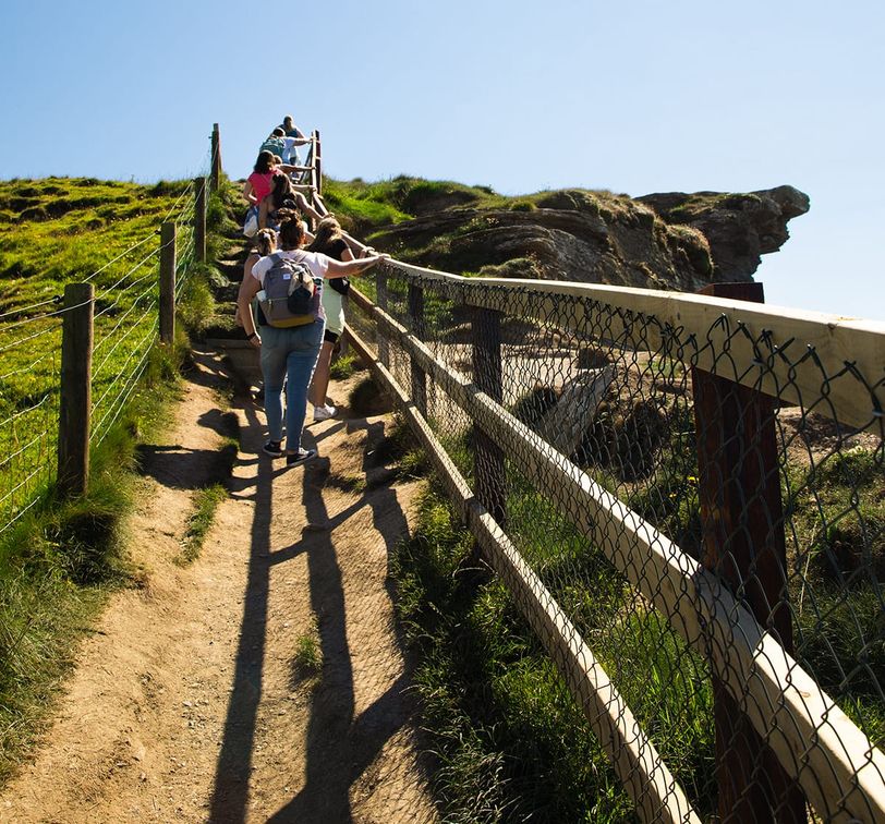 People walking up a narrow dirt path on a sunny day, flanked by grassy hills and a wooden fence, leading to a rocky cliff edge.