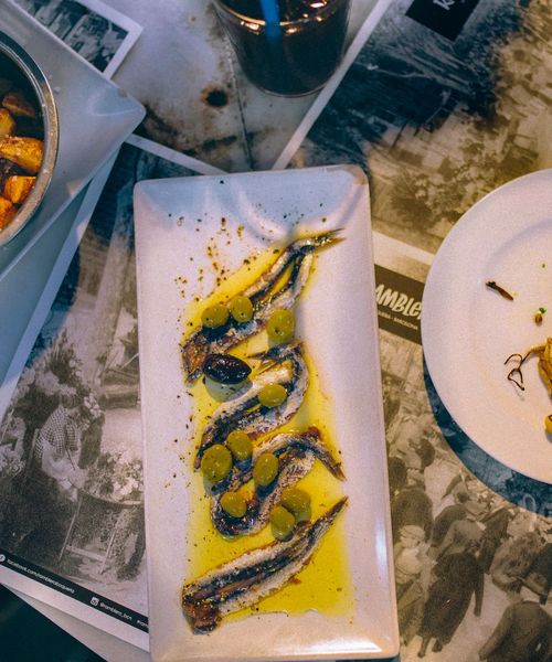 Plate of marinated anchovies with olives and olive oil, accompanied by roasted potatoes and a partially eaten dish on a table.