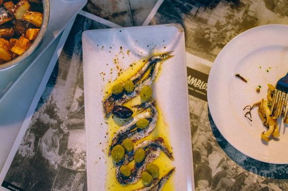 Plate of marinated anchovies with olives and olive oil, accompanied by roasted potatoes and a partially eaten dish on a table.