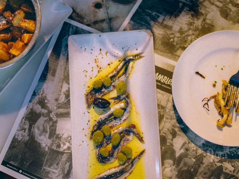 Plate of marinated anchovies with olives and olive oil, accompanied by roasted potatoes and a partially eaten dish on a table.