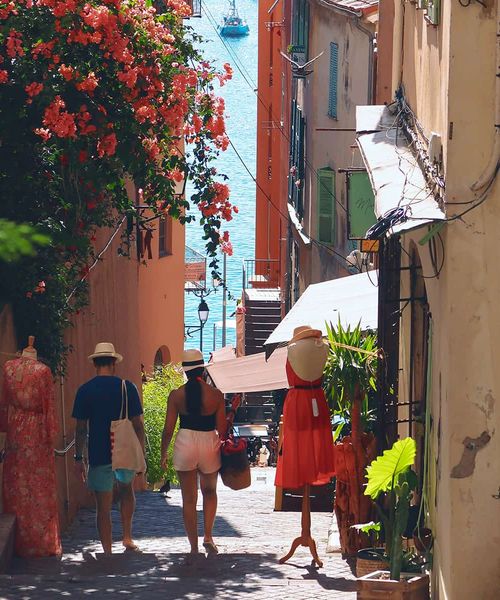 A couple walks down a narrow, colorful street lined with flowers, mannequins, and shops, towards the sea in the background.