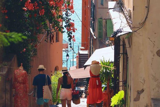 A couple walks down a narrow, colorful street lined with flowers, mannequins, and shops, towards the sea in the background.