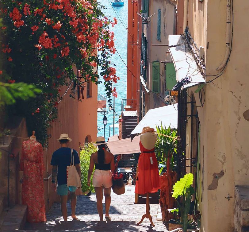 A couple walks down a narrow, colorful street lined with flowers, mannequins, and shops, towards the sea in the background.