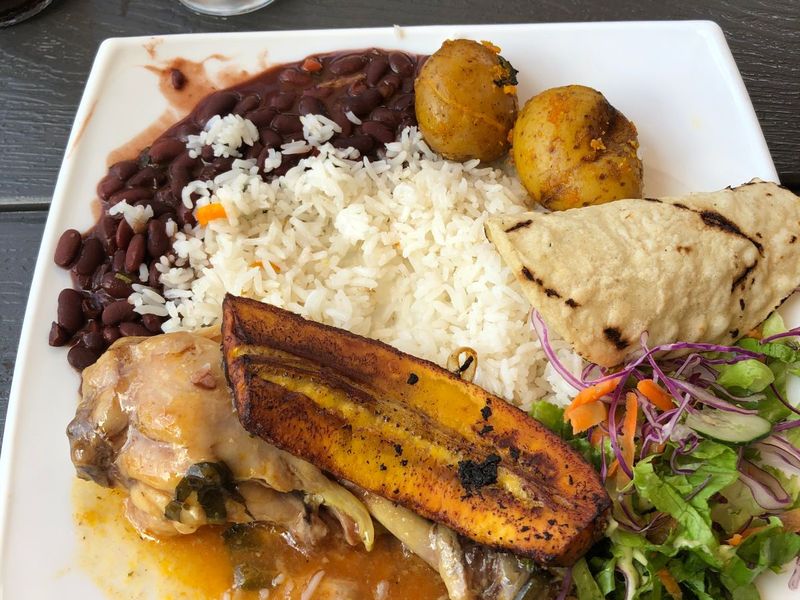 A plate with rice, black beans, stewed chicken, fried plantain, potatoes, tortilla, and salad with shredded carrots and purple cabbage.