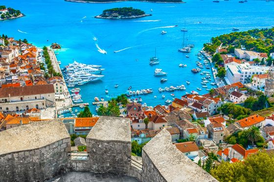 Aerial view of a coastal town with red-roofed buildings, boats in a vibrant blue harbor, and small lush islands in the background.