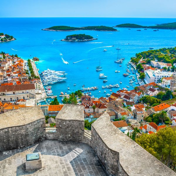 Aerial view of a coastal town with red-roofed buildings, boats in a vibrant blue harbor, and small lush islands in the background.