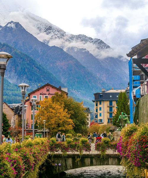 Scenic view of a charming town with colorful buildings, lush flowers on a bridge, and snow-capped mountains in the background.
