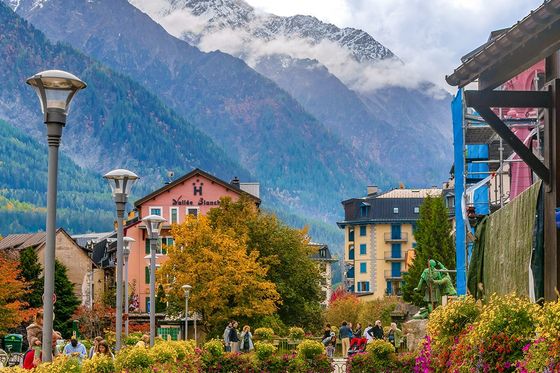 Scenic view of a charming town with colorful buildings, lush flowers on a bridge, and snow-capped mountains in the background.