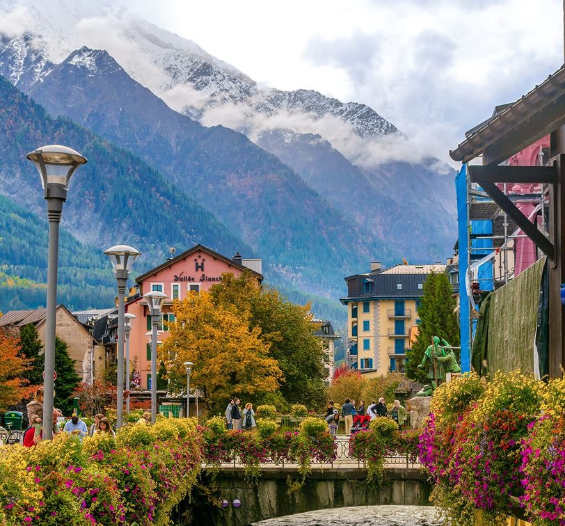 Scenic view of a charming town with colorful buildings, lush flowers on a bridge, and snow-capped mountains in the background.