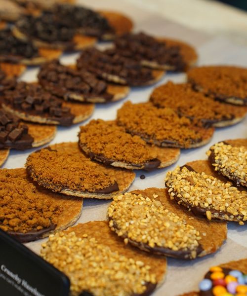 A display of cookies with various toppings, including chocolate pieces, crushed nuts, and cookie crumbs, arranged neatly on parchment paper.