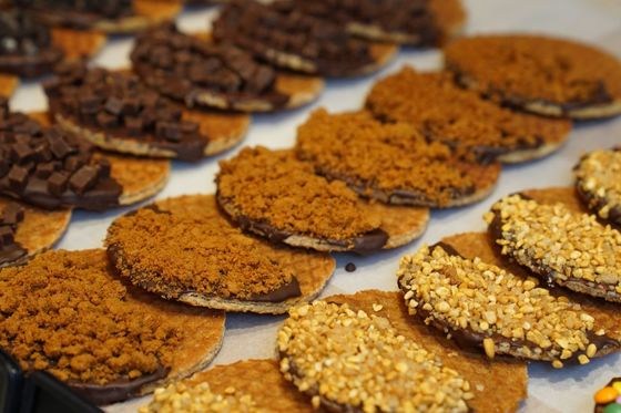 A display of cookies with various toppings, including chocolate pieces, crushed nuts, and cookie crumbs, arranged neatly on parchment paper.