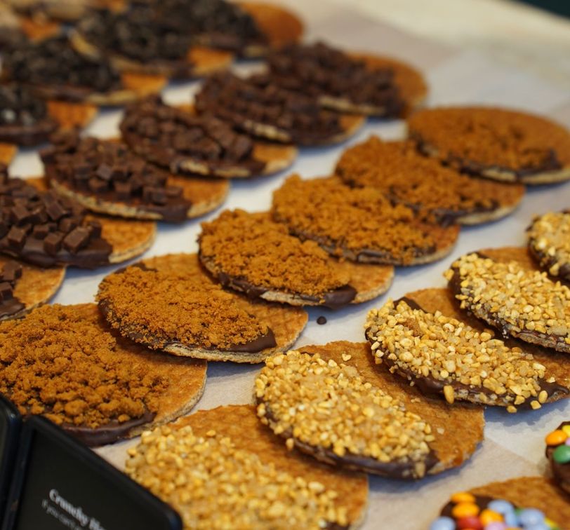 A display of cookies with various toppings, including chocolate pieces, crushed nuts, and cookie crumbs, arranged neatly on parchment paper.