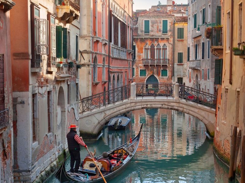 A gondolier navigates a narrow canal in Venice, flanked by historic buildings, with a small arched bridge overhead and soft sunlight illuminating the scene.