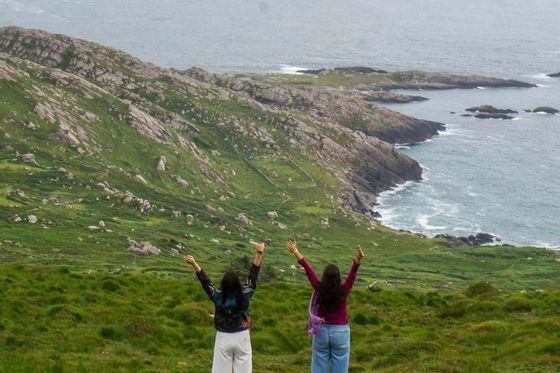 Two people with raised arms stand on a grassy hill overlooking a rocky coastline and the ocean.