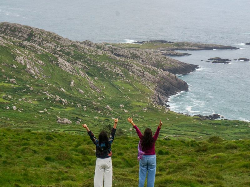 Two people with raised arms stand on a grassy hill overlooking a rocky coastline and the ocean.