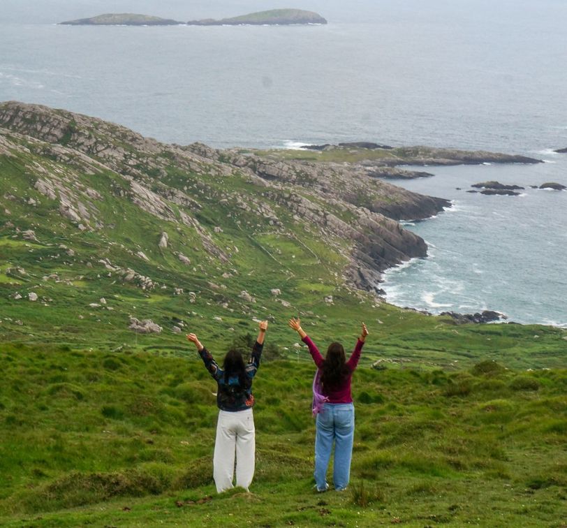 Two people with raised arms stand on a grassy hill overlooking a rocky coastline and the ocean.