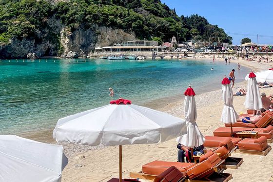 Sunny beach scene with loungers and umbrellas along a sandy shore. Clear turquoise water and a lush green hill in the background.