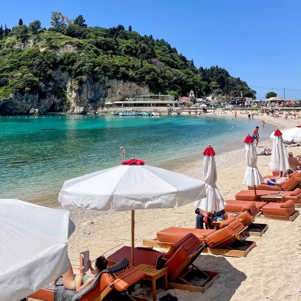 Sunny beach scene with loungers and umbrellas along a sandy shore. Clear turquoise water and a lush green hill in the background.