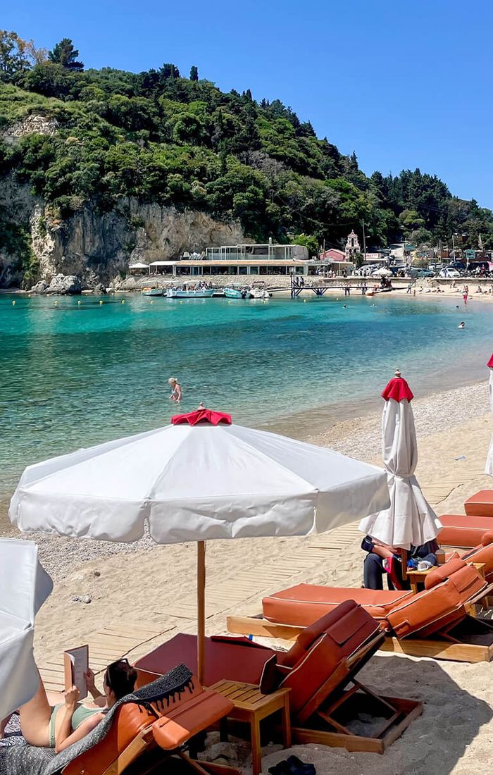 Sunny beach scene with loungers and umbrellas along a sandy shore. Clear turquoise water and a lush green hill in the background.