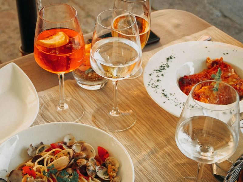 A wooden table with plates of seafood pasta and lasagna, accompanied by glasses of orange and clear drinks, elegantly arranged for a meal.