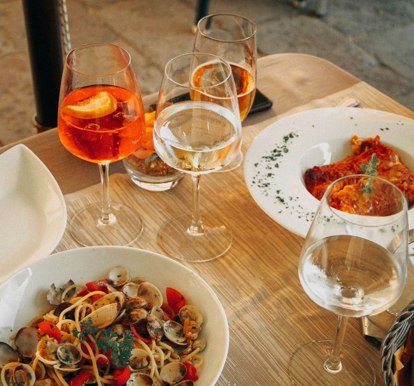 A wooden table with plates of seafood pasta and lasagna, accompanied by glasses of orange and clear drinks, elegantly arranged for a meal.