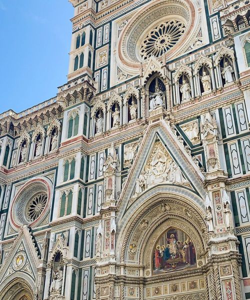 Intricate details of a cathedral facade, featuring ornate sculptures, circular stained glass windows, and architectural embellishments under a clear blue sky.
