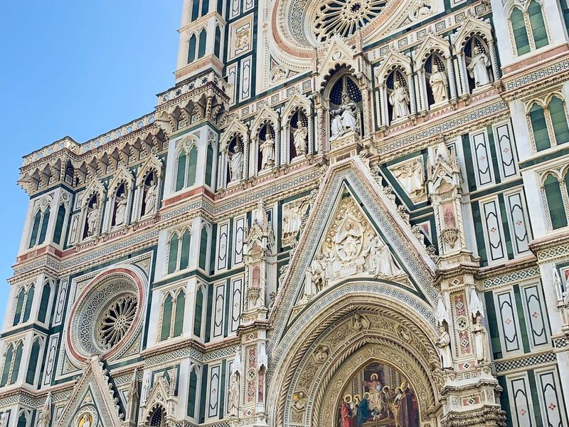 Intricate details of a cathedral facade, featuring ornate sculptures, circular stained glass windows, and architectural embellishments under a clear blue sky.