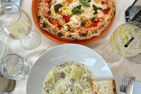 A table with a pizza topped with tomatoes, cheese, and herbs. Below, a creamy pasta dish with mushrooms, served with a slice of bread, surrounded by drink glasses.