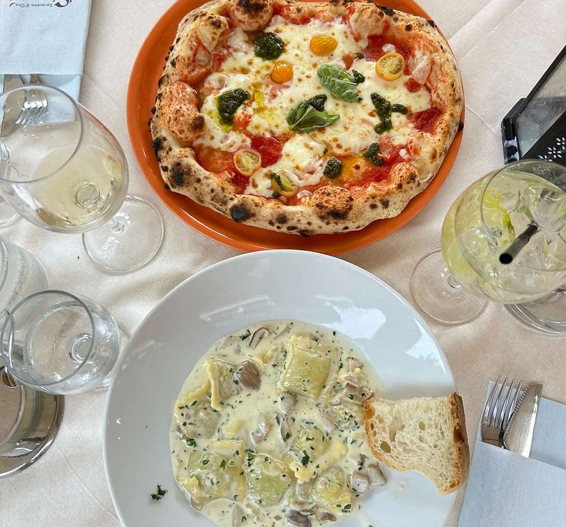 A table with a pizza topped with tomatoes and basil, a pasta dish with creamy sauce and mushrooms, wine glasses, and a slice of bread.