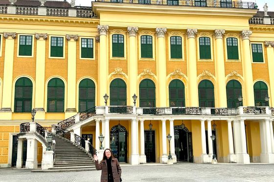 Person in a dark coat standing in front of a large, ornate yellow building with green window shutters and tall columns.