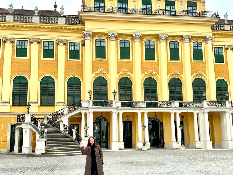 Person in a dark coat standing in front of a large, ornate yellow building with green window shutters and tall columns.