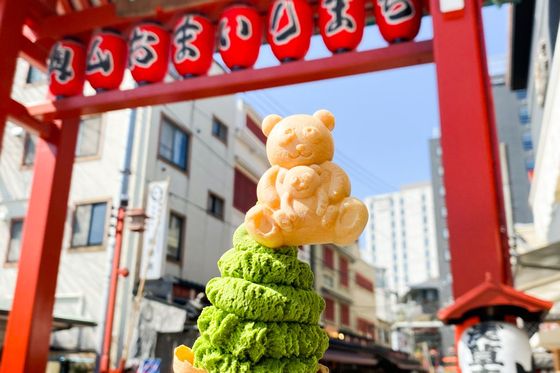 Matcha ice cream in a decorative cone topped with a bear wafer, held in front of a red Japanese gate with hanging lanterns.
