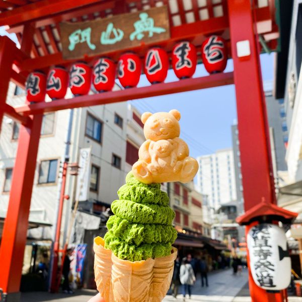 Matcha ice cream in a decorative cone topped with a bear wafer, held in front of a red Japanese gate with hanging lanterns.